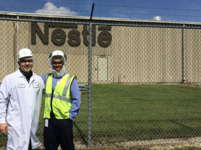 Two people in protective gear standing in front of an industrial facility.